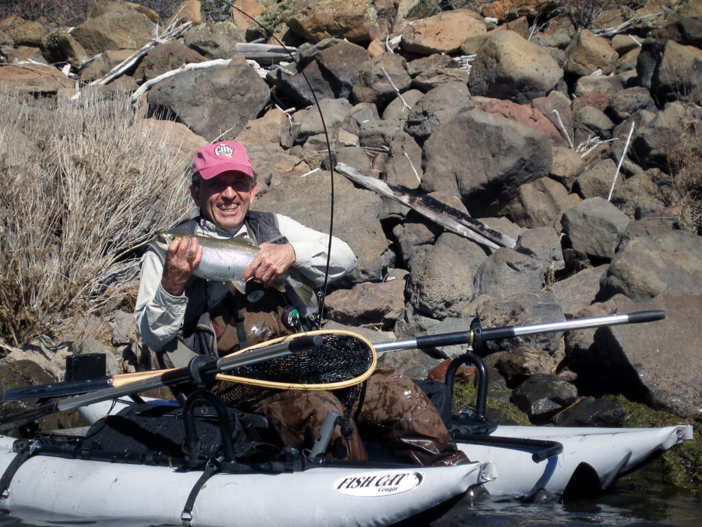 A man in a personal fishing pontoon boat holding a rainbow trout along a rocky shoreline
